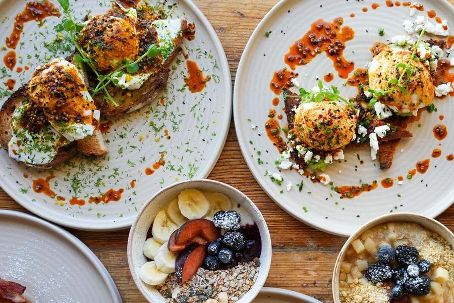 Two plates of poached eggs on toast with chilli butter, beside bowls of granola and porridge, on a wooden table